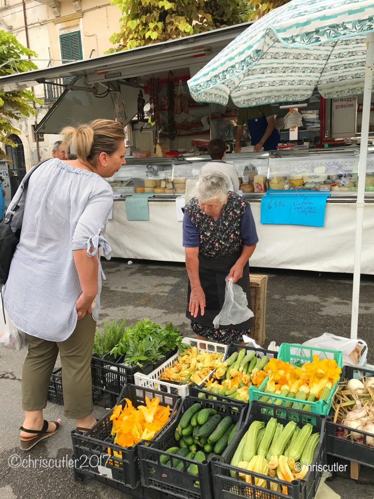 woman buying vegetables from woman vendor; fish truck in background