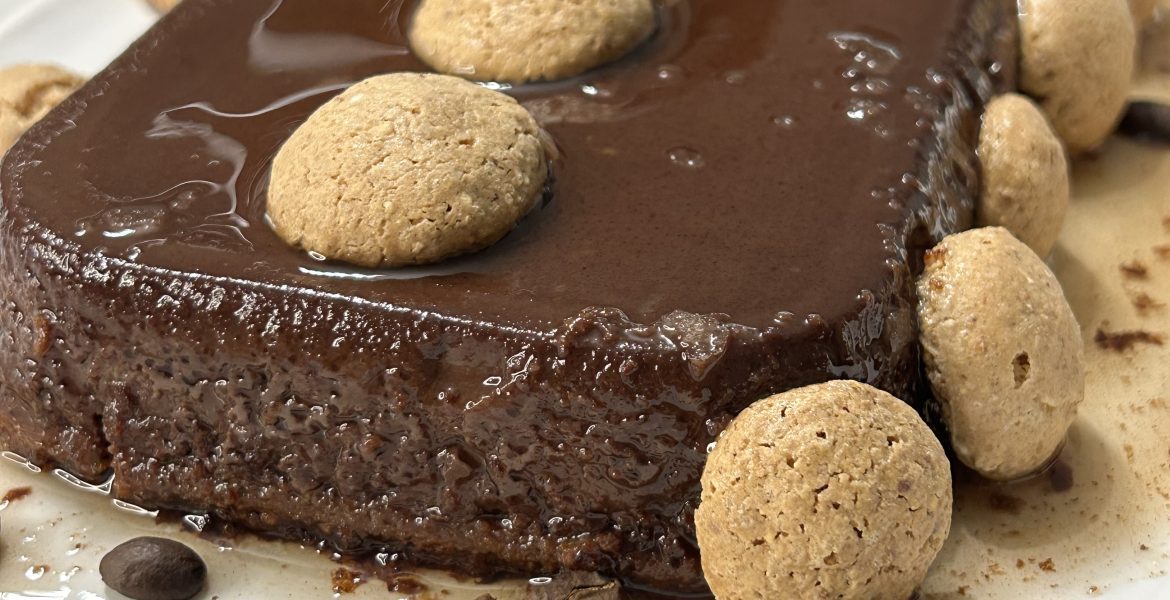 A slice of decadent Italian chocolate flan (bonet) with a thin caramel layer on top, garnished with amaretti cookies and coffee beans.