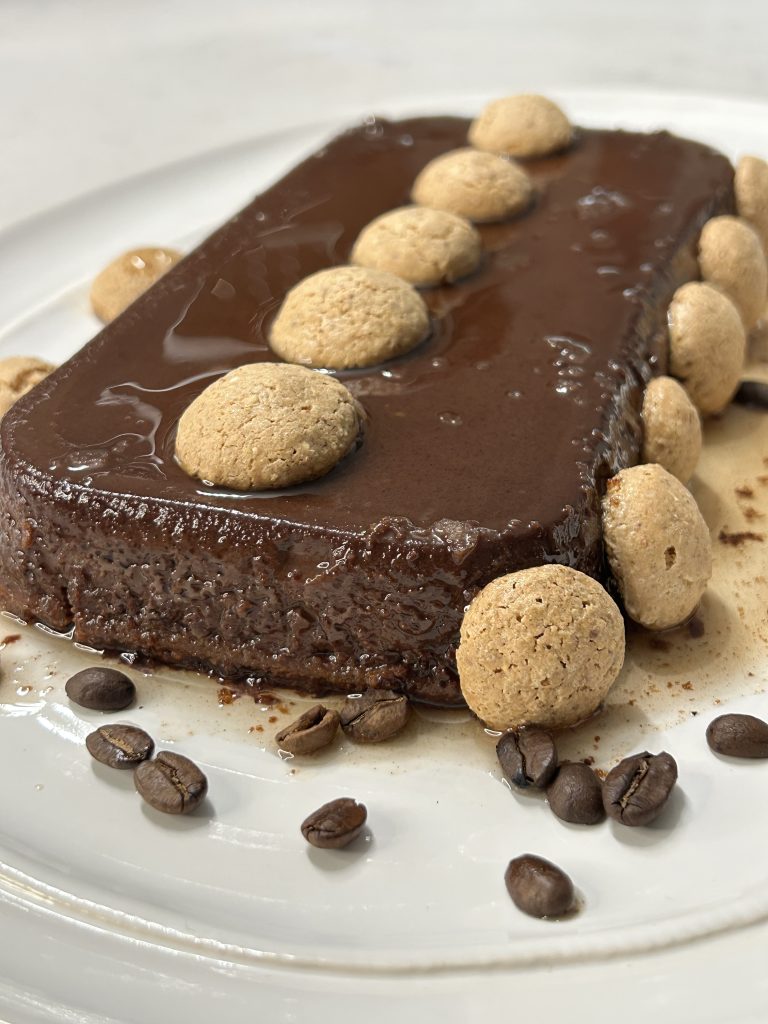 A slice of decadent Italian chocolate flan (bonet) with a thin caramel layer on top, garnished with amaretti cookies and coffee beans.