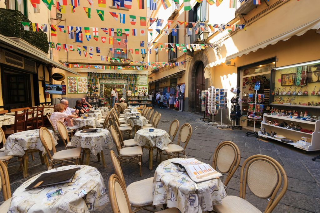 A cobblestone alley in Sorrento, Italy, with outdoor dining and shops. The shop Sorrento È is visible in the background, showcasing the town's famous appeal for both food and shopping.