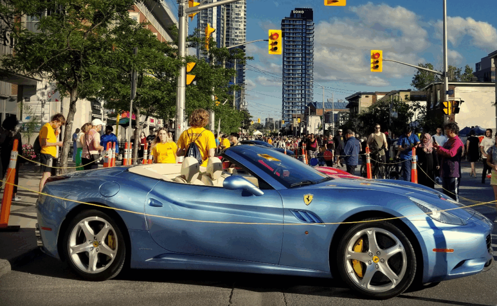A light blue Ferrari convertible sports car parked on Preston Street amid a crowd of people during the Ottawa Ferrari Festival.