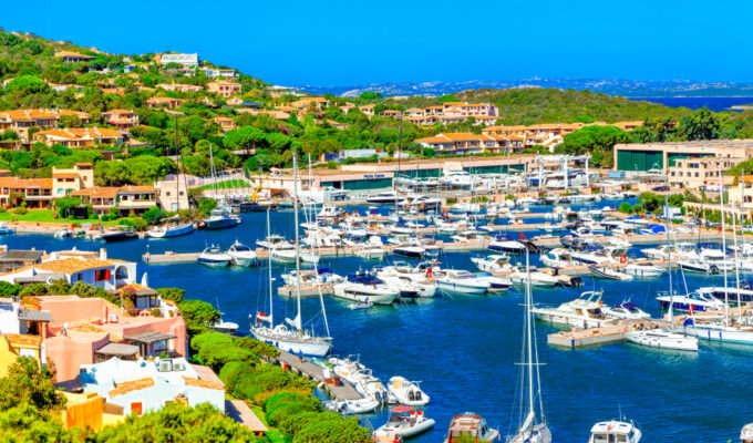 Panoramic view of Porto Cervo marina on the Costa Smeralda in Sardegna, with luxury yachts, turquoise water, and Mediterranean hillside homes under a bright blue sky.