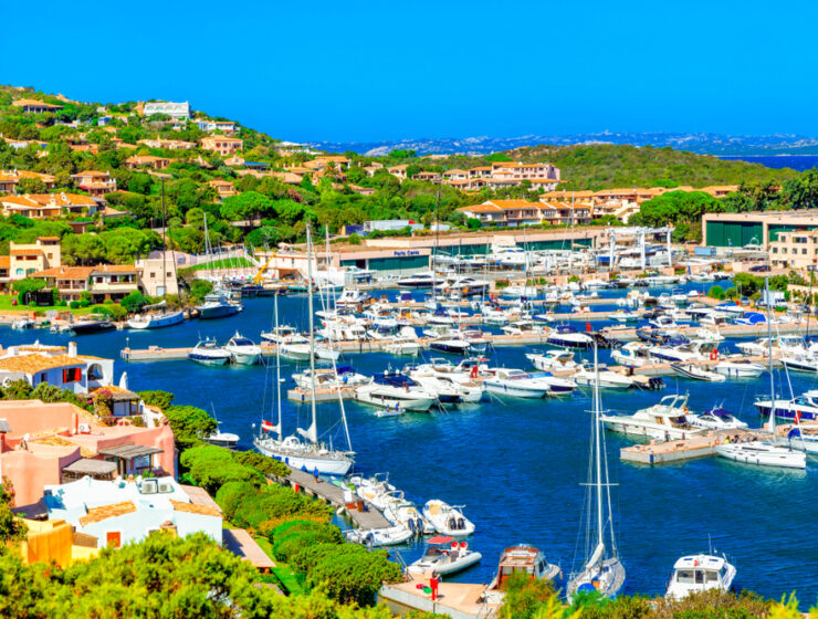 Panoramic view of Porto Cervo marina on the Costa Smeralda in Sardegna, with luxury yachts, turquoise water, and Mediterranean hillside homes under a bright blue sky.