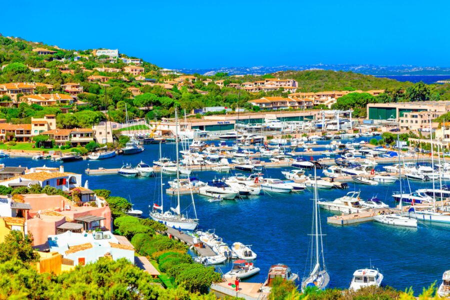 Panoramic view of Porto Cervo marina on the Costa Smeralda in Sardegna, with luxury yachts, turquoise water, and Mediterranean hillside homes under a bright blue sky.