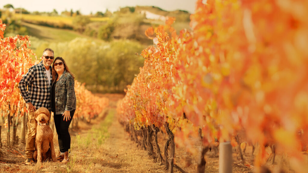 Sandy and Phil Ferretti smiling together in a Tuscan vineyard during autumn, surrounded by rows of golden and orange grapevine leaves.