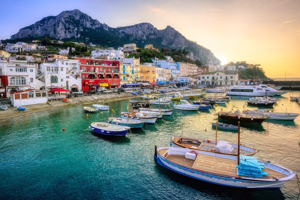Colorful waterfront buildings and fishing boats in Marina Grande on the island of Capri, with mountains rising behind the harbor at sunset.