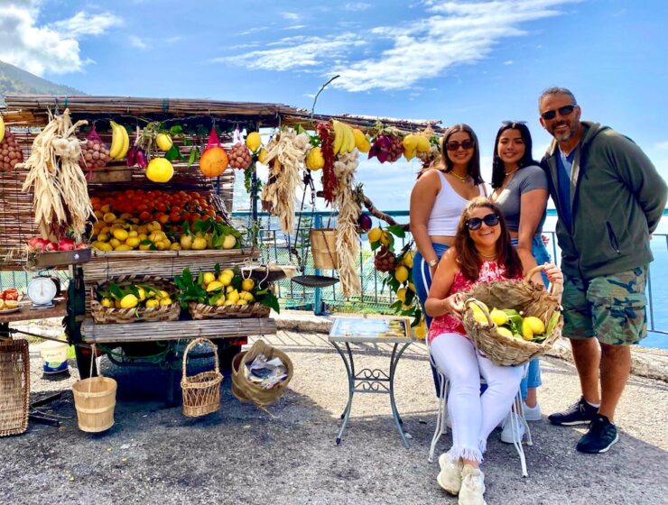 Sandy and Phil with two travelers beside a colorful roadside fruit stand displaying fresh lemons and other produce along the Amalfi Coast.