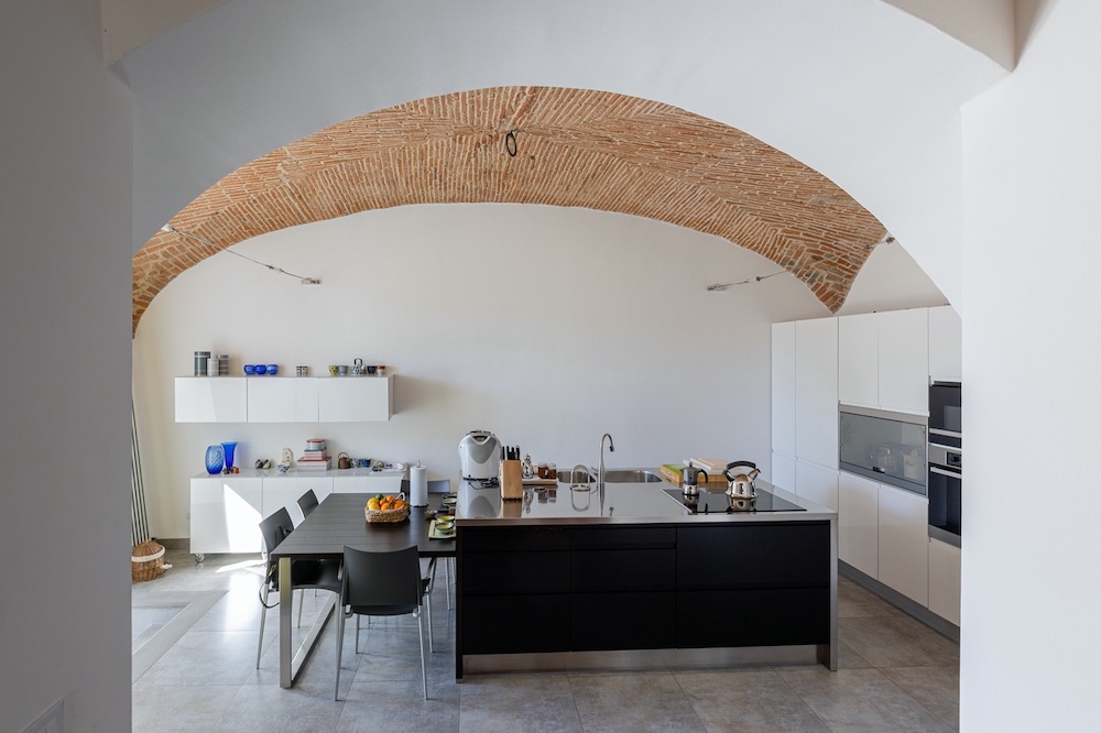Contemporary kitchen with a large exposed brick arch, black island and white cabinetry inside a restored historic abbey residence in Mugello, Tuscany.