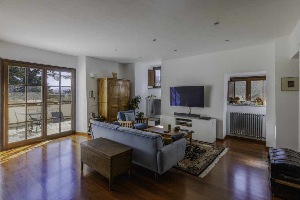 Sunlit living room with wooden floors, blue seating and glass doors opening onto a terrace at Badia di Santa Maria a Vigesimo in Tuscany.