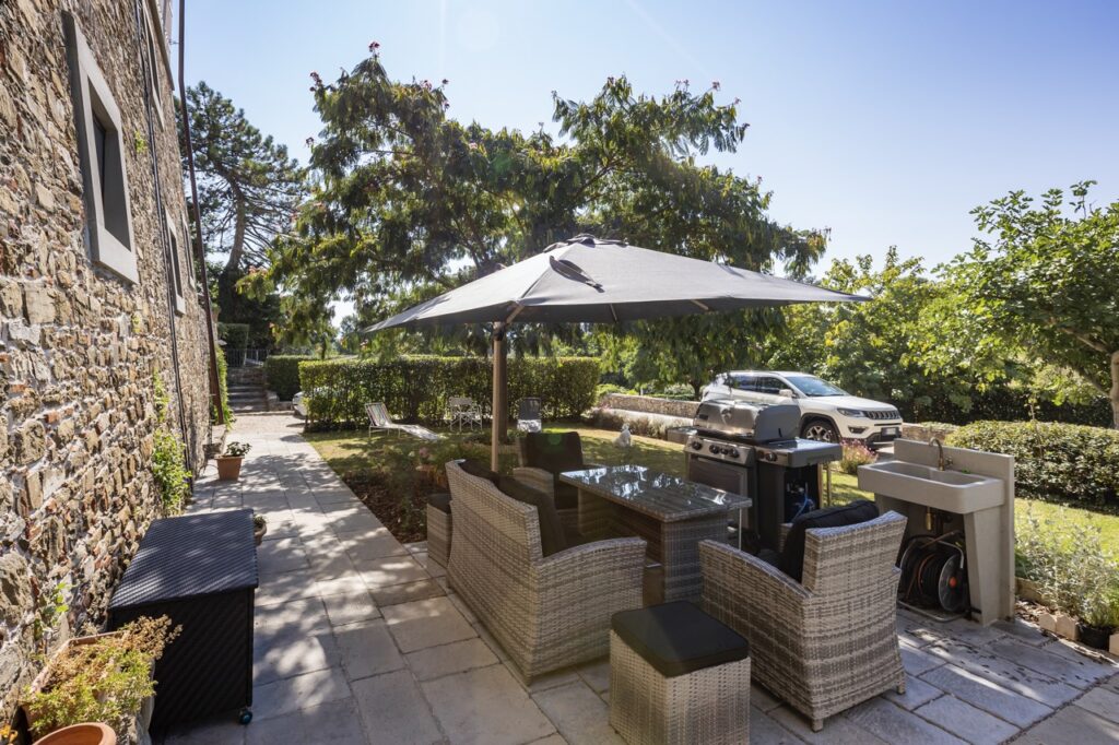 Private garden patio with outdoor dining furniture, barbecue grill and mature trees beside the stone walls of a restored abbey residence in Mugello, Tuscany.