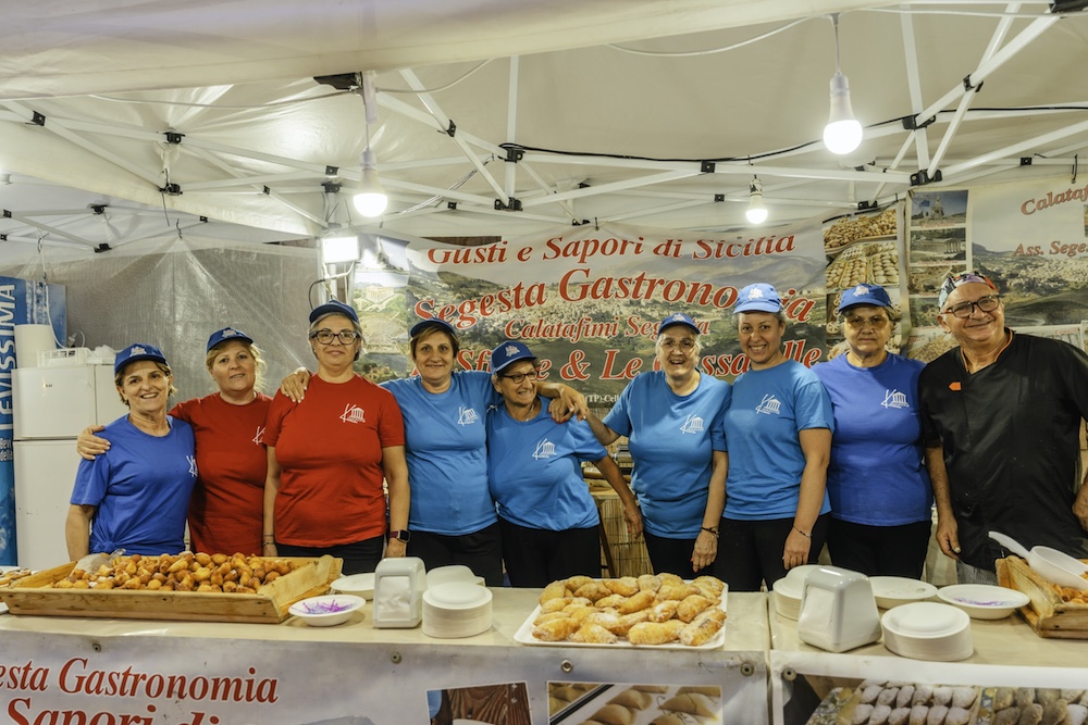 Festival volunteers and food vendors behind a counter displaying traditional Sicilian pastries during the Sagra della Busiata in Salemi.
