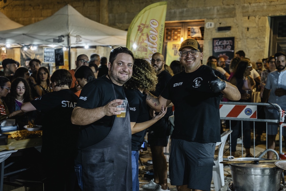 Two smiling food stall workers surrounded by visitors during the evening Sagra della Busiata festival in Salemi, Sicily.
