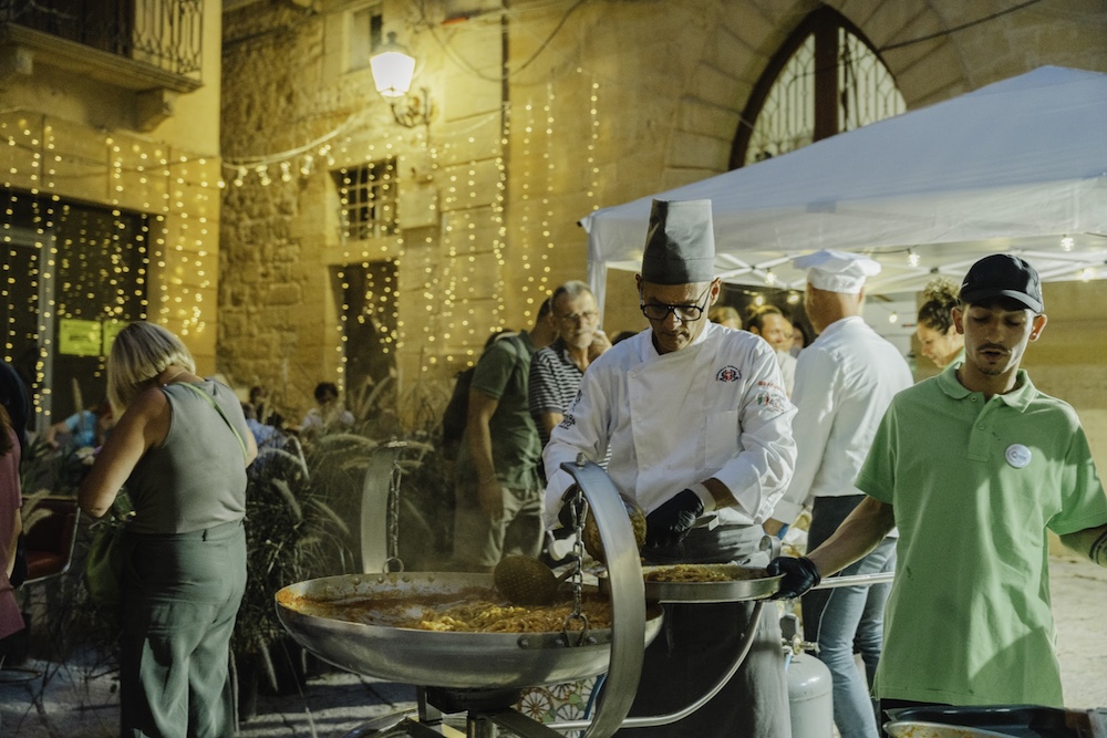 Chef preparing Sicilian pasta in a large cooking pan at an outdoor food station during Salemi's Sagra della Busiata festival.
